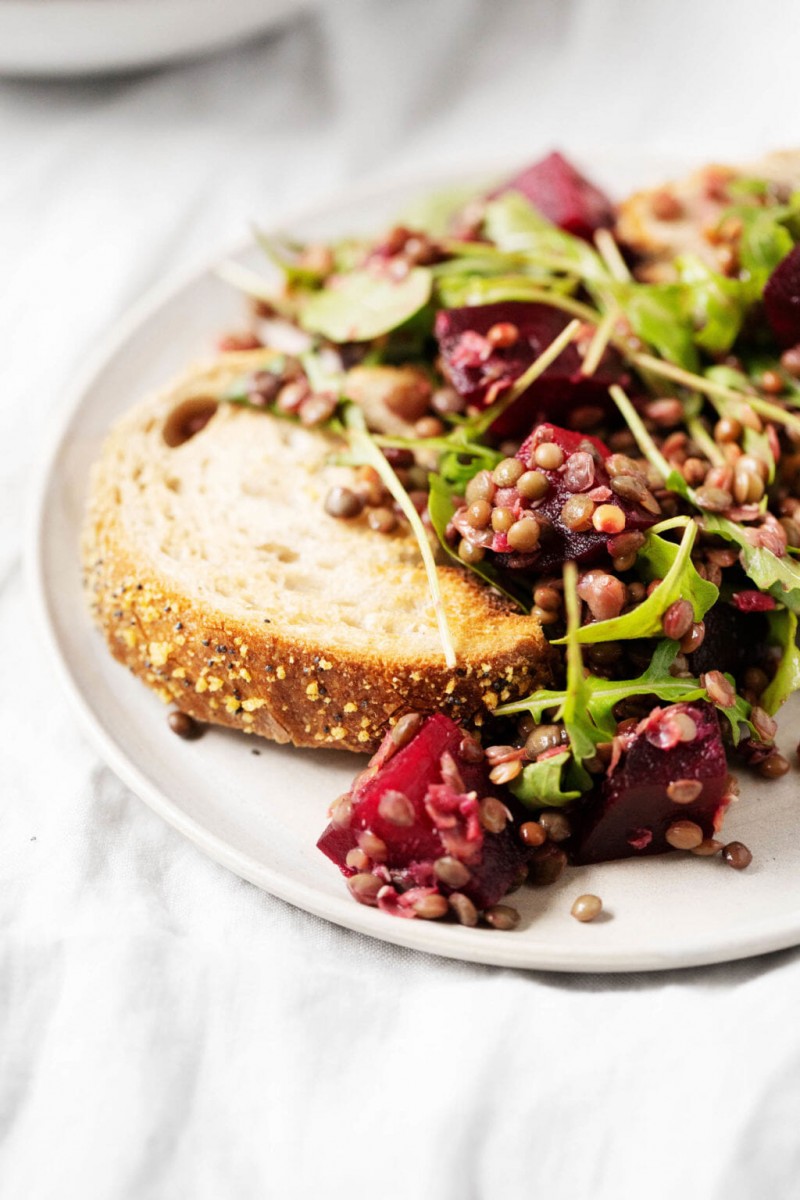 A zoomed in image of a slice of seeded bread, which has been piled with the leftovers of a lentil beet salad.