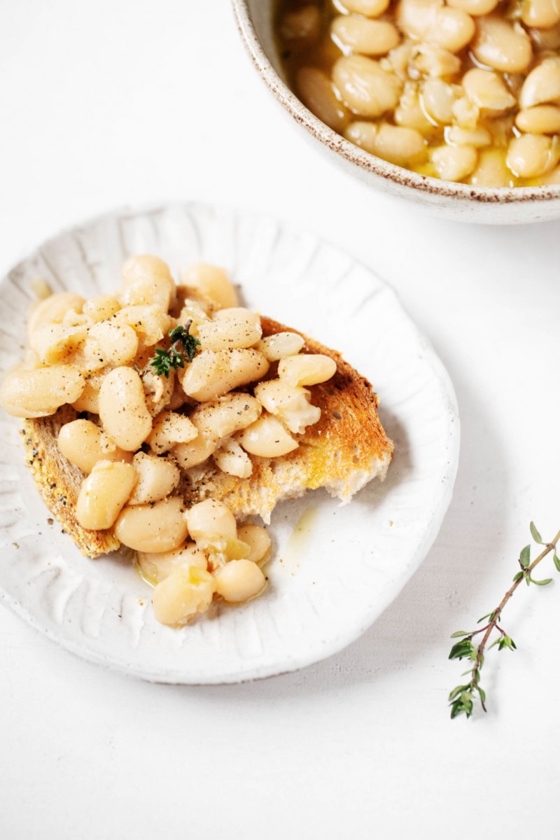 Two small serving plates hold toast that has been covered in braised, brothy white beans. A sprig of thyme rests on a nearby surface.