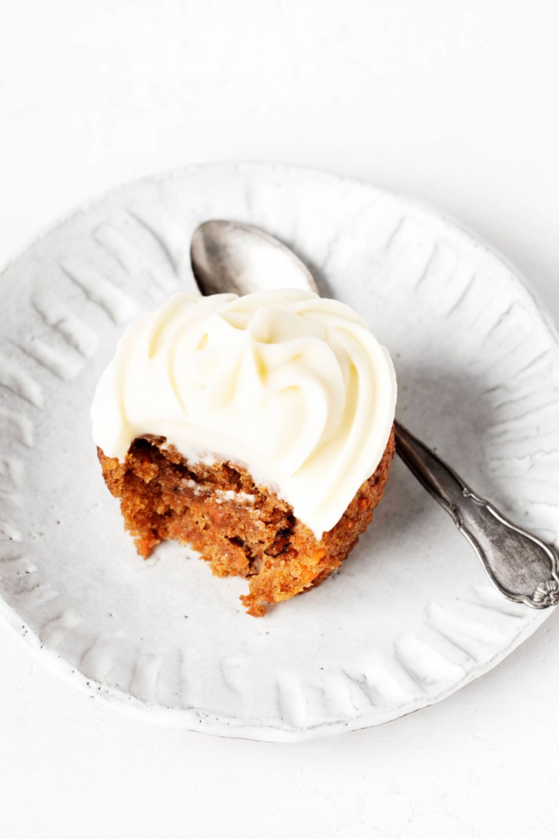 A single vegan carrot cake cupcake rests on a small, ceramic white plate. There's a small spoon beside the cupcake.