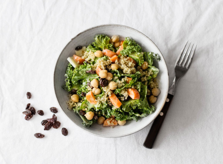 A white ceramic bowl holds a carrot raisin kale salad. A fork rests nearby.