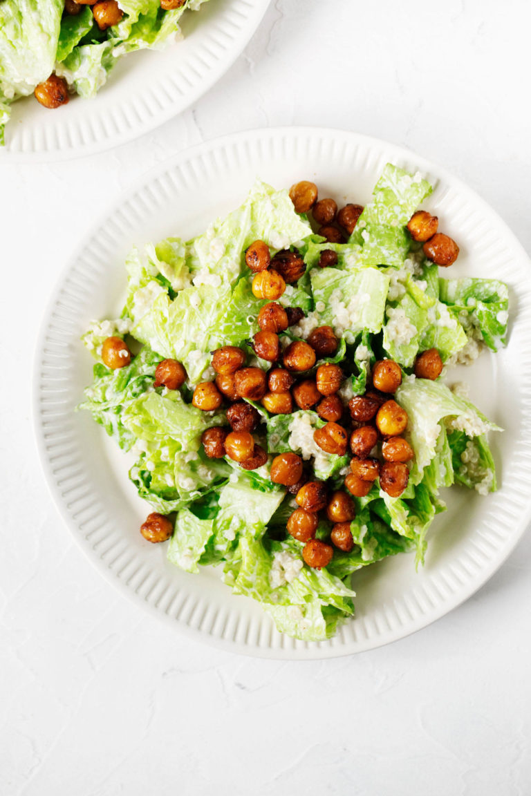 Two white, fluted ceramic plates have been piled with a vegan quinoa chickpea caesar salad. The plates are resting on a white background.