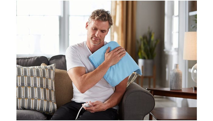 A man sitting on a couch presses a light blue heating pad onto his left shoulder with his right hand.