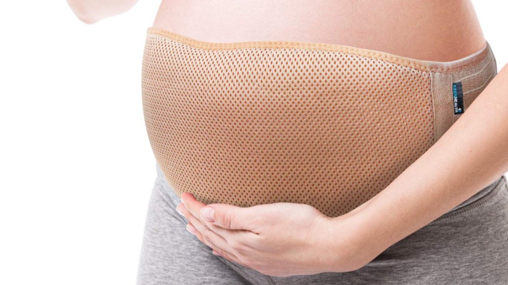 A pregnant woman wearing a beige back support belt with her hand resting on her lower belly.