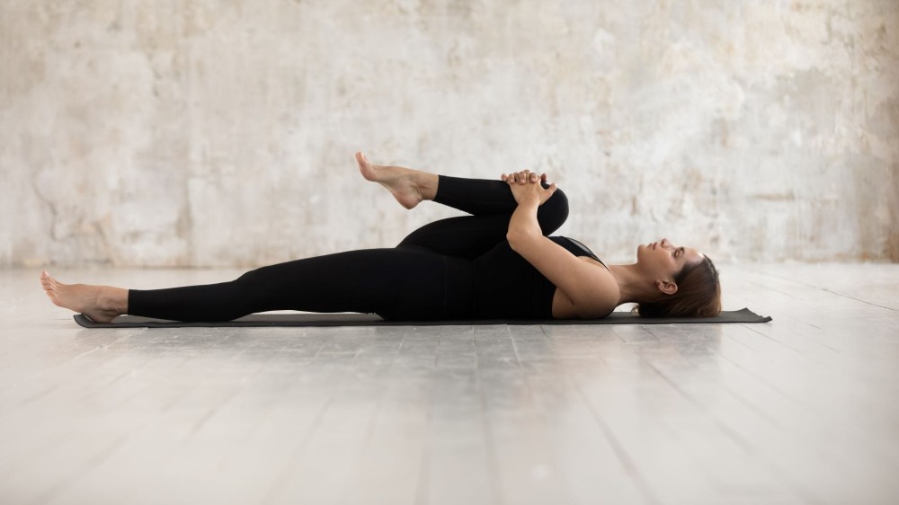 A woman lying on her back on a yoga mat clutching one knee to her chest.