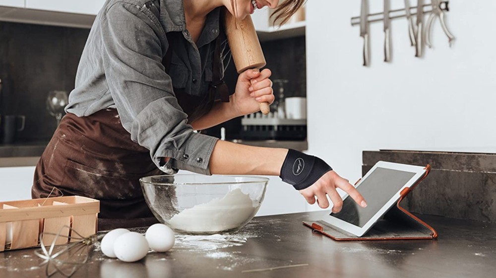 A woman surrounded by baking tools and ingredients wears a black wrist brace while looking at recipe.