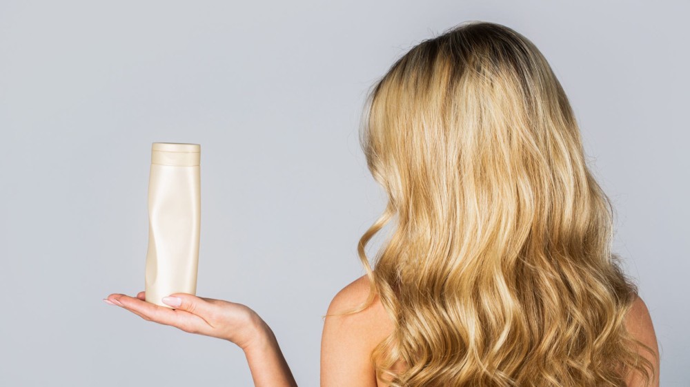 A blonde woman with shiny pretty hair holds a bottle of shampoo out.