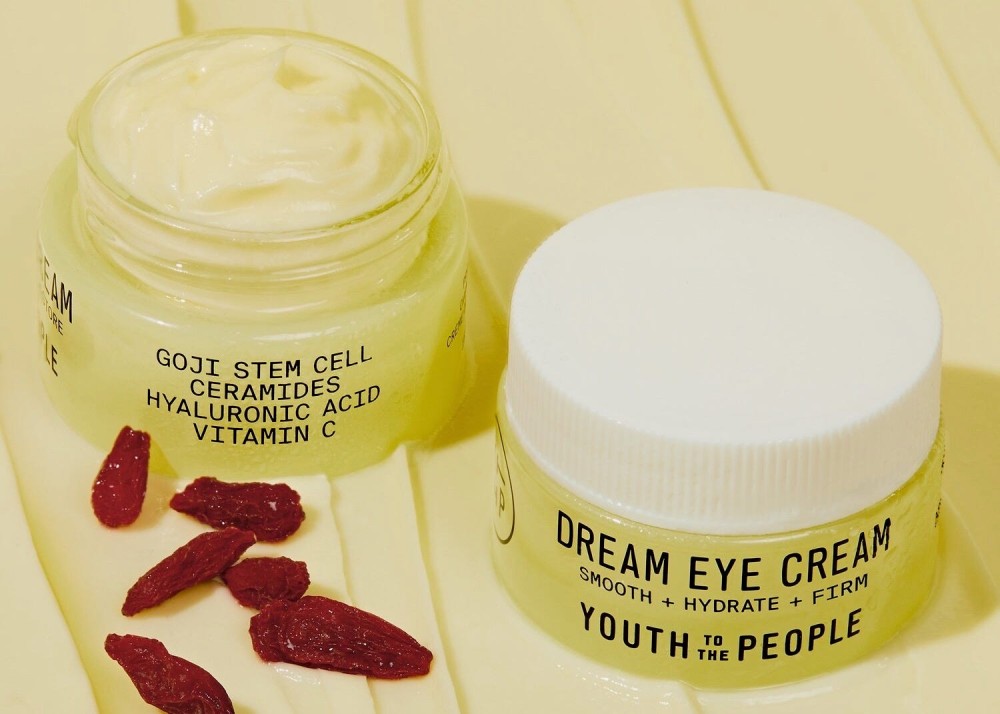 Two containers of eye cream, one open and one closed, next to dried goji berries.