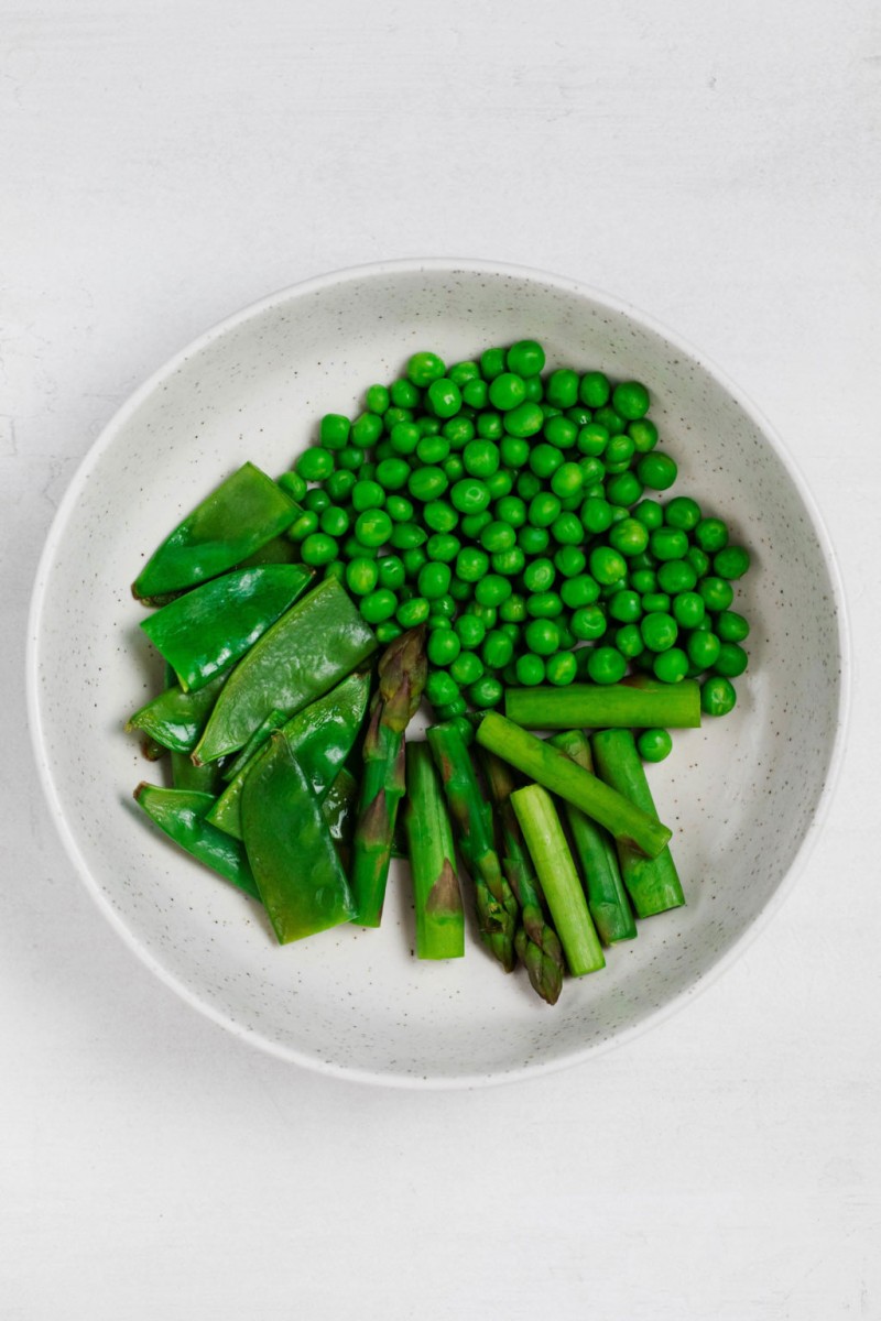 A round, white bowl is filled with various types of green vegetables. It rests on a white surface.