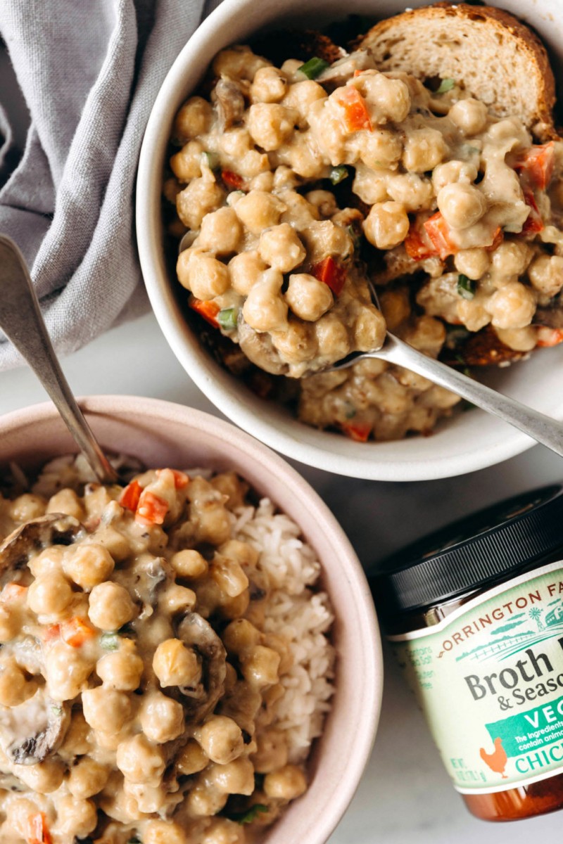 Two white bowls hold a dish of chickpeas à la King. A container of bouillon powder and gray napkin lie nearby.