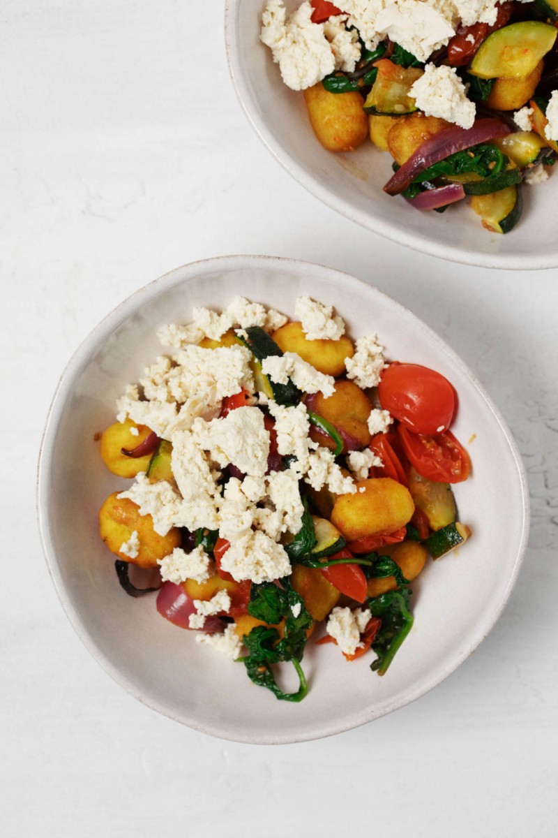 Two bowls of pillowy, roasted potato dumplings are served in white bowls with roasted tomatoes and zucchini. The bowls are topped with plant-based cheese.