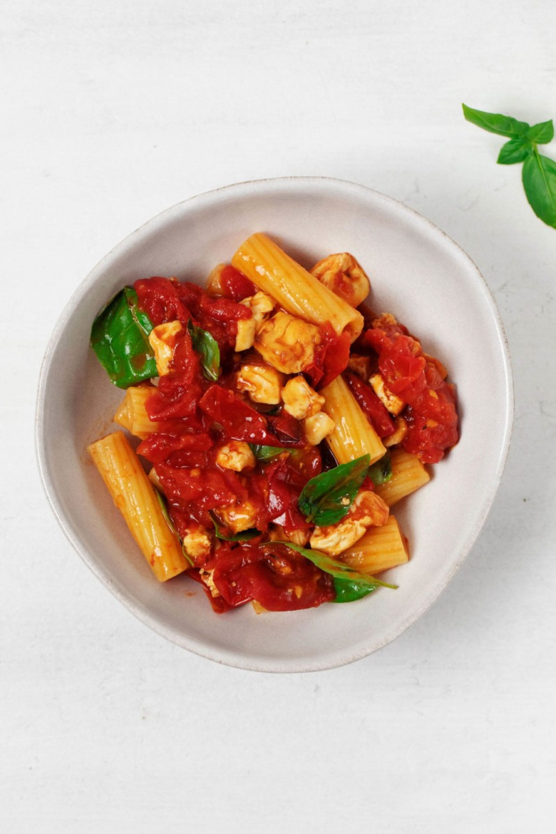 An overhead image of a round bowl of vegan baked feta pasta, prepared with tofu. The bowl rests on a white surface.