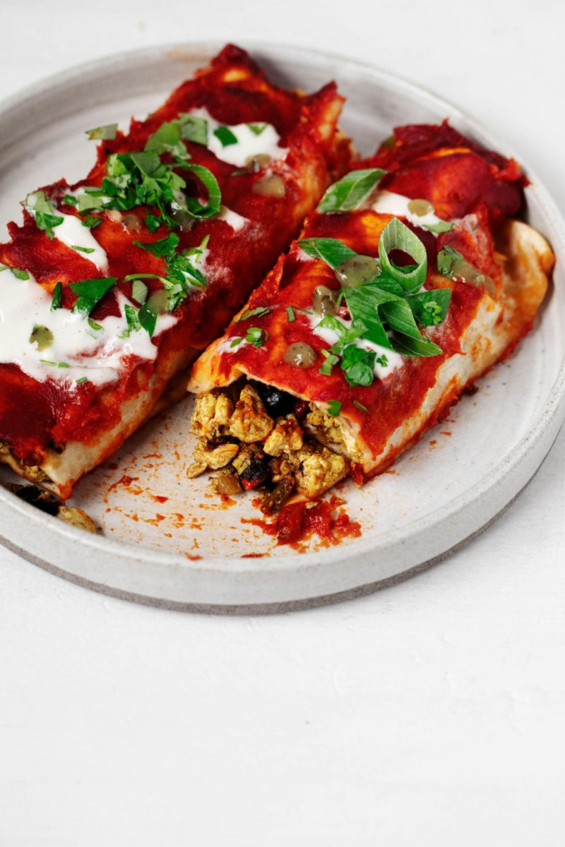 A white, rimmed plate holds two vegan enchiladas that have been filled with tofu scramble and black beans.