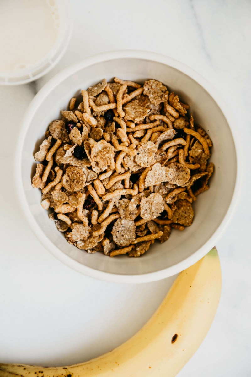 A bowl of cereal rests next to a banana and a cup of milk on a white marble surface.