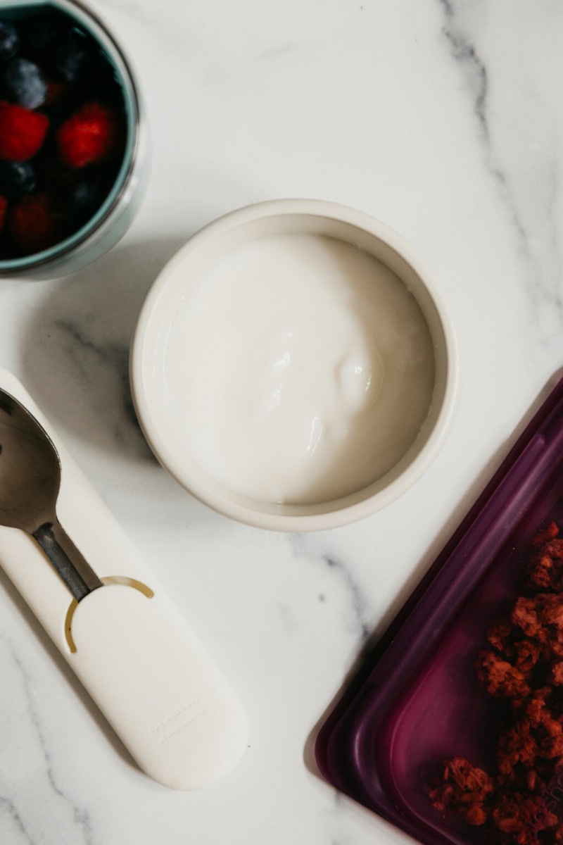 A container of yogurt has been packed to go. It's accompanied by a silicone bag of granola and some berries.