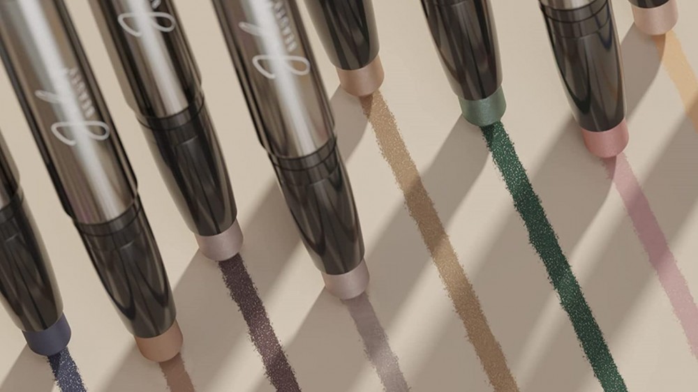 Many colors of eyeshadow pencils drawing a rainbow of shimmering makeup lines on white paper.