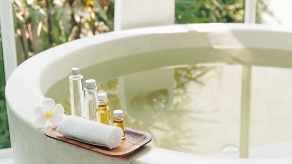 Nnatural organic bath products on a wooden tray on the edge of a big bathtub.