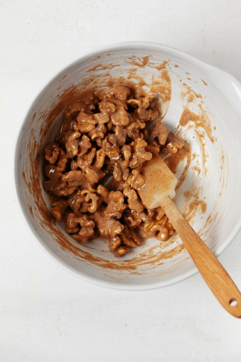 A white mixing bowl is filled with walnut halves and pieces that are being glazed.