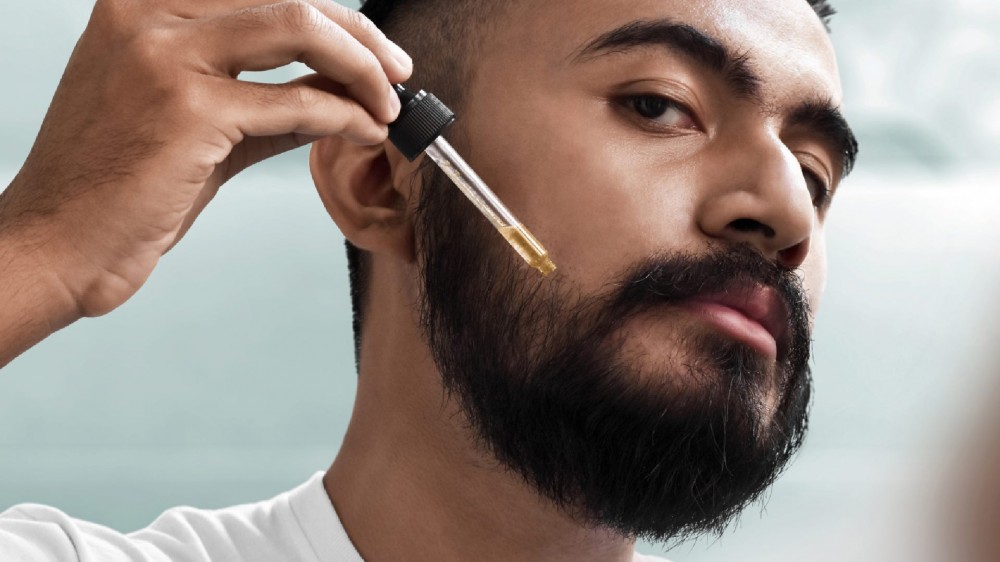 Bearded man holding a pipette with beard oil in it over his beard.