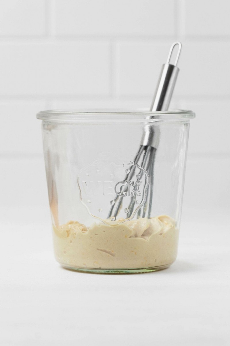 Tahini is in a glass mason jar, along with a small, metal whisk. The jar rests on a white surface.