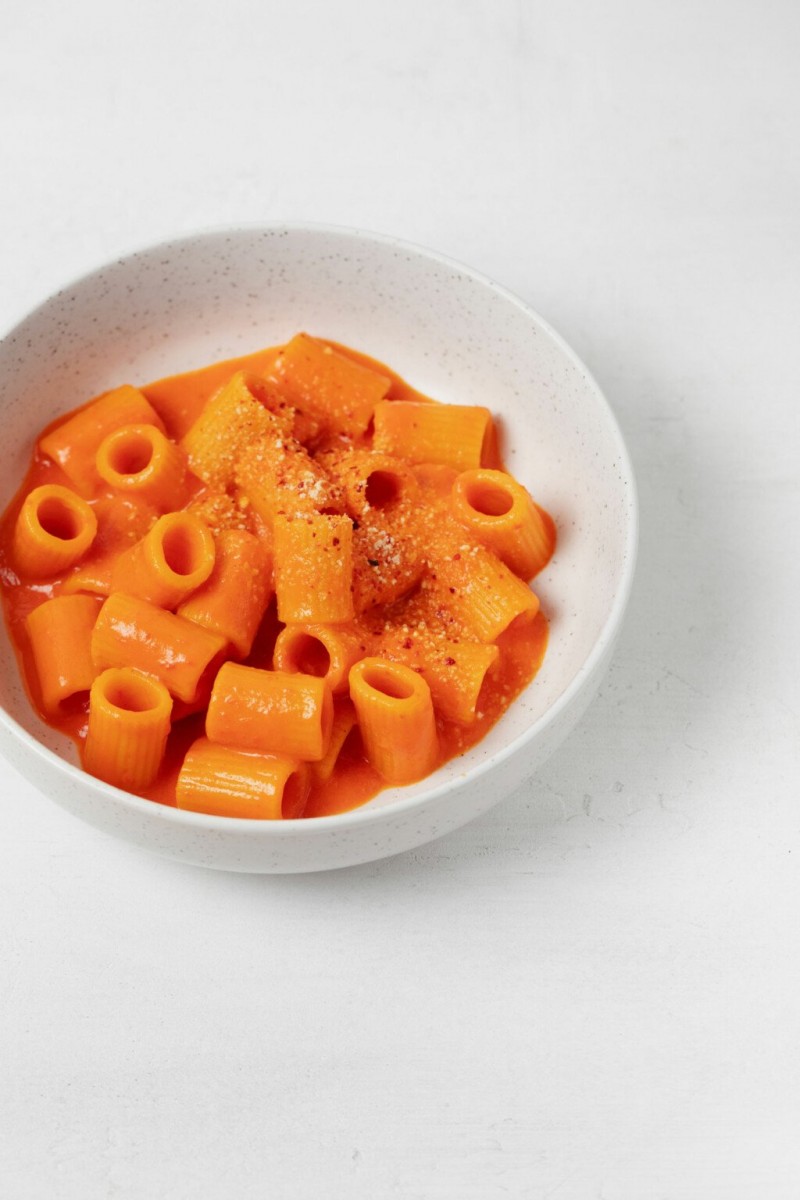 A bowl of creamy red pepper pasta has been topped with a vegan "cheese" topping. It's plated in a white bowl.