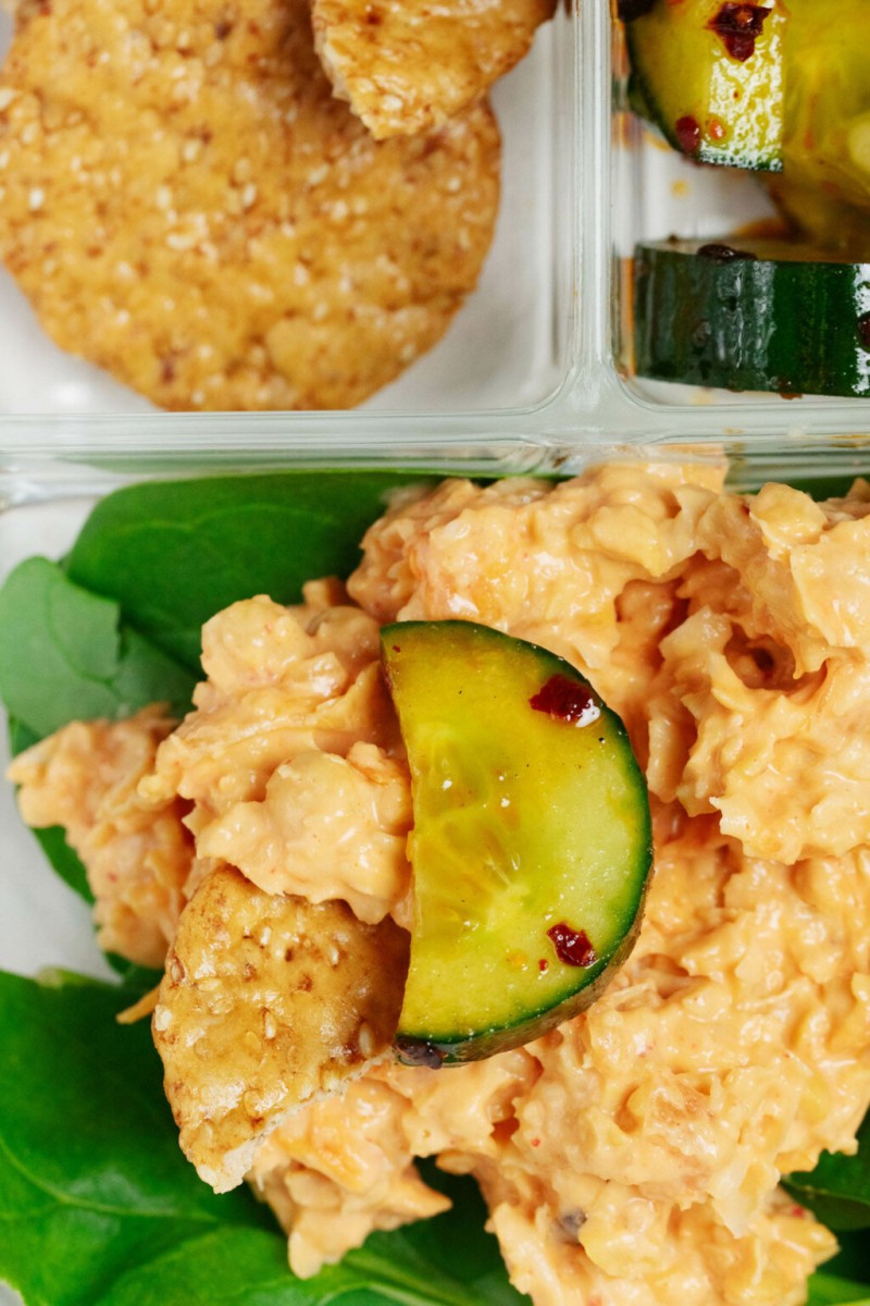 An overhead image of smashed chickpea salad with kimchi, which is served in a lunch box over greens, along with a small cucumber slice.