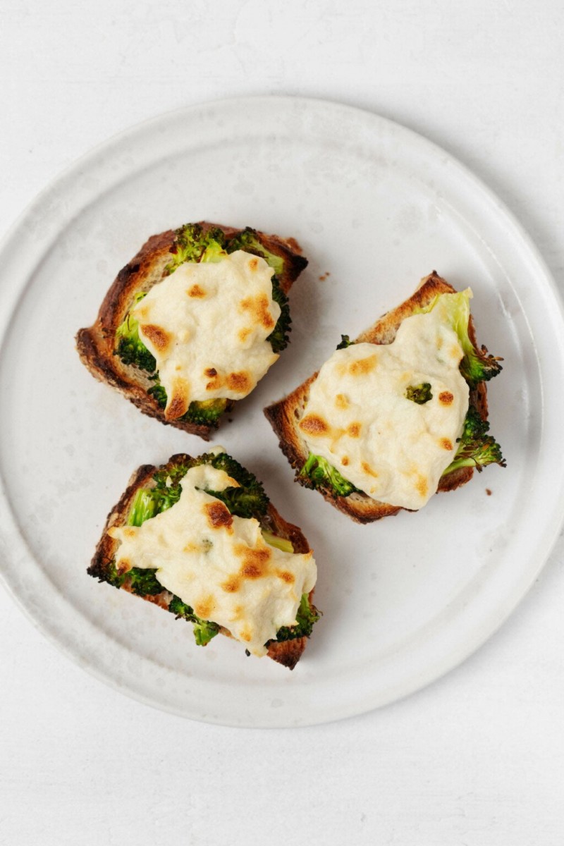 An overhead image of three plant-based broccoli melts on whole grain bread. They rest on a round, white plate.