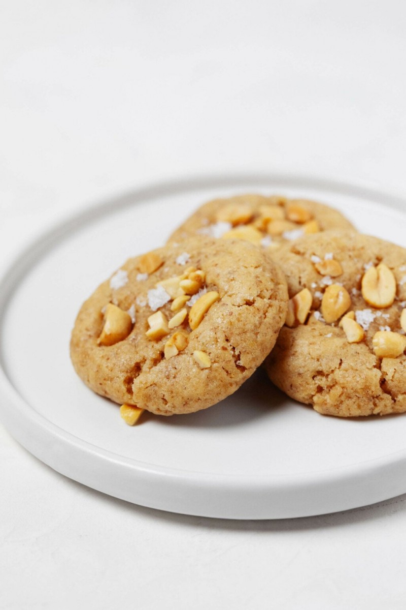 A round, rimmed white plate holds three vegan peanut cookies, each topped with chopped peanuts and flaky salt.