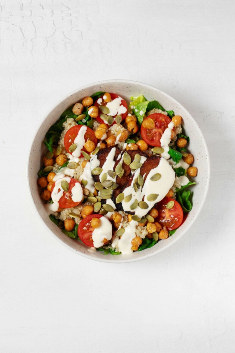 A large, white bowl is filled with vegetables, roasted chickpeas, quinoa, and a creamy ranch dressing. The bowl is topped with pepitas.