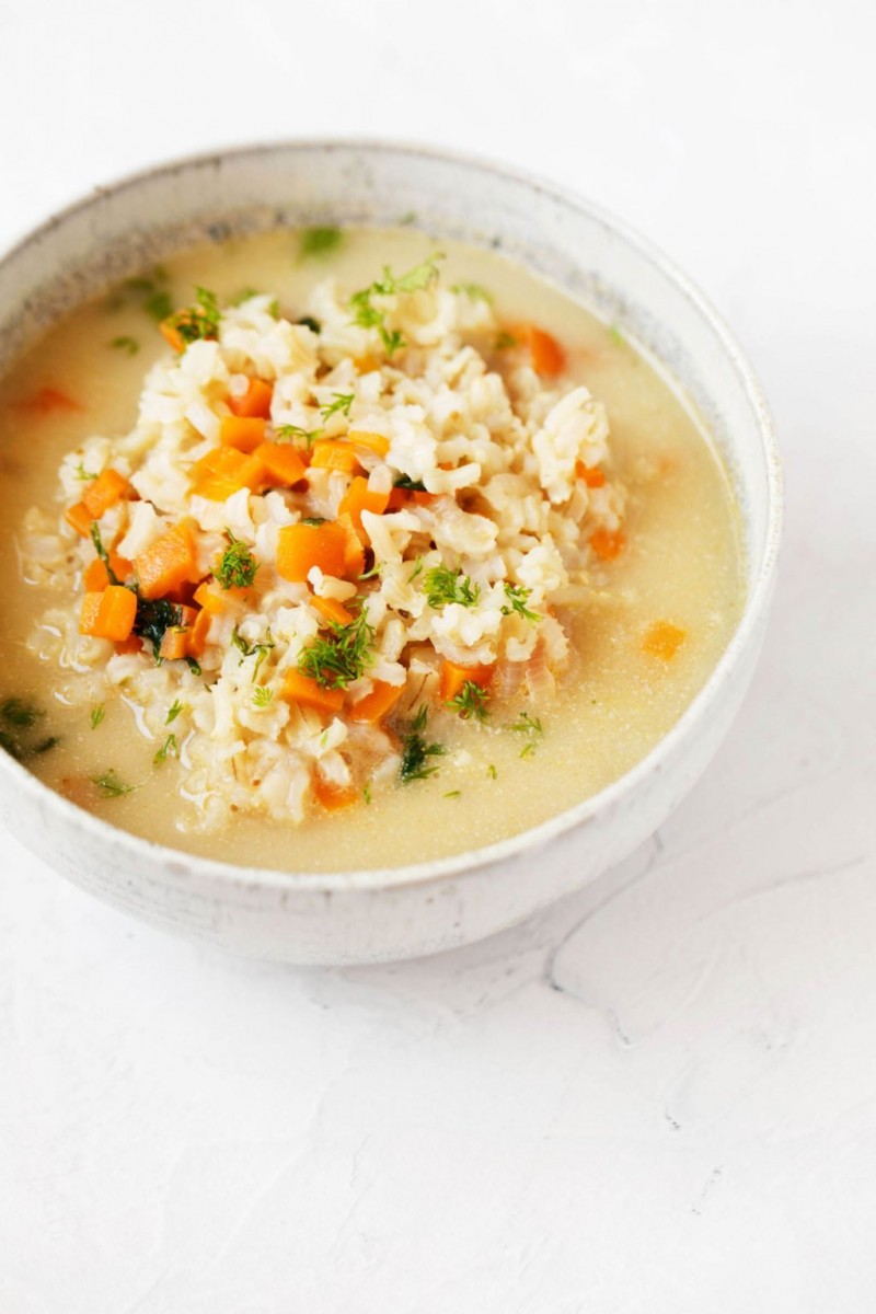 A white-rimmed, ceramic bowl holds a brothy mixture of rice, carrots, celery, and herbs. It rests on a white surface.