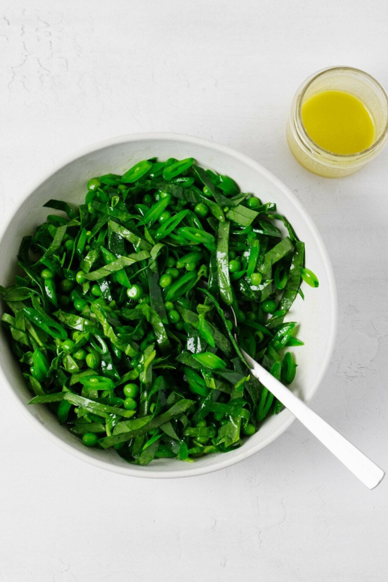 An overhead image of a pea salad, made with bright green shelled peas and thinly sliced snow peas. A jar of vinaigrette rests on a white surface nearby.